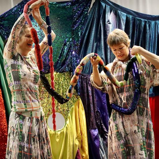 To kvindelige skuespillere i en farverig kulisser med forskellige ruller af farvet stof.