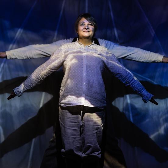 To mennesker i en teateropsætning med lys, den ene står forrest med armene spredt ud til siderne, den anden står bagerst, så man kun kan se vedkommendes arme. 